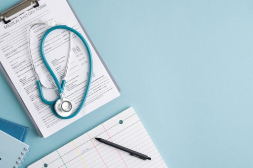 Top view of medical history form in clipboard, folded stethoscope, notebooks and pen on blank card w